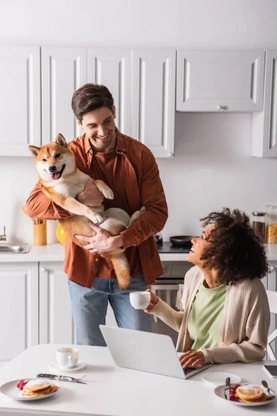 Счастливая межрасовая пара глядя друг на друга рядом шиба Ину собака и ноутбук на кухне — стоковое фото