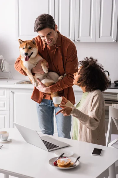 African american woman pointing at laptop near smiling boyfriend with shiba inu dog — Stock Photo