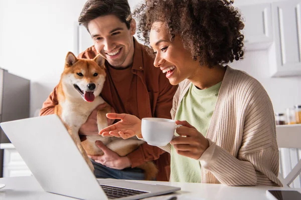 Sorridente donna afro-americana in possesso di tazza vicino dispositivi e fidanzato con shiba inu a casa — Foto stock