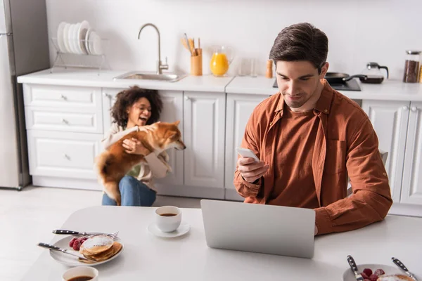 Homme utilisant smartphone et ordinateur portable près du petit déjeuner et petite amie afro-américaine avec shiba inu dans la cuisine — Photo de stock