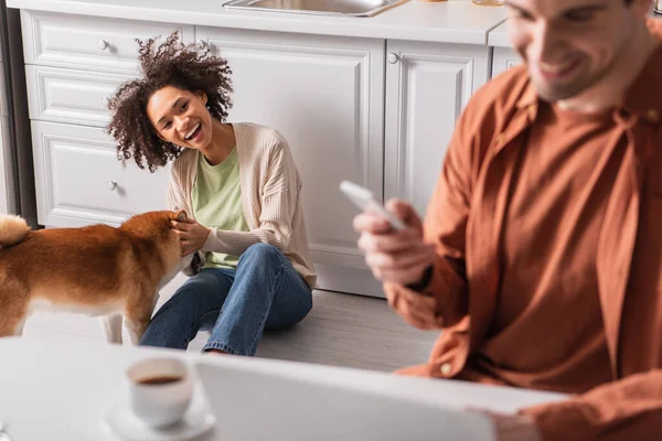 Sonriente mujer afroamericana jugando con shiba inu cerca de novio borroso con smartphone en la cocina - foto de stock