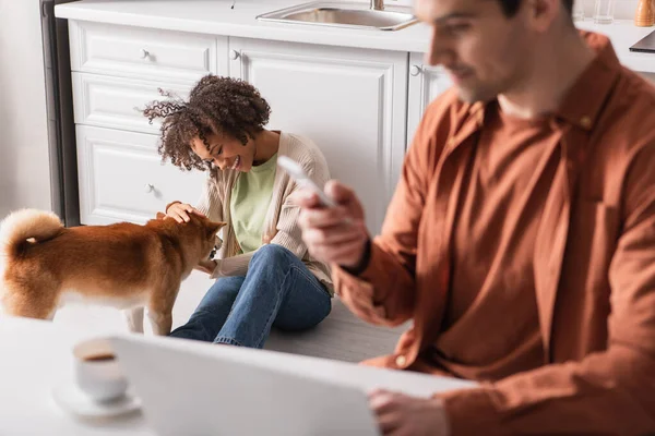 Joven mujer afroamericana jugando con shiba inu cerca de novio con dispositivos en la cocina - foto de stock