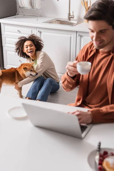 Positivo africano americano mujer jugando con shiba inu cerca borrosa novio con taza usando laptop en cocina - foto de stock