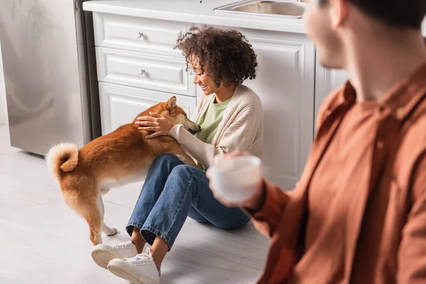 Young african american woman playing with shiba inu dog near blurred boyfriend with cup in kitchen — Stock Photo