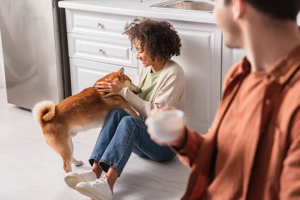 Joven afroamericana mujer jugando con shiba inu perro cerca borrosa novio con taza en cocina - foto de stock