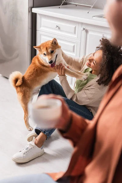 Sorridente donna africana americana che gioca con shiba inu vicino uomo sfocato con tazza in cucina — Foto stock