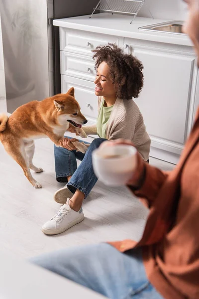 Afroamerikanerin spielt mit Shiba Inu in der Küche in der Nähe ihres Freundes mit Tasse — Stockfoto