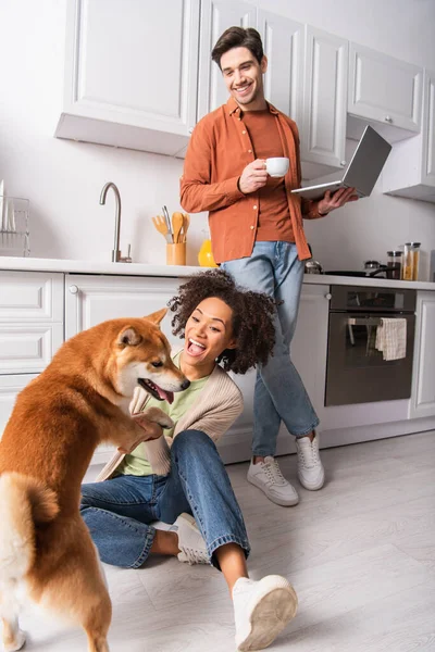 Afro-américaine femme jouer avec shiba inu près petit ami avec ordinateur portable et café dans la cuisine — Stock Photo