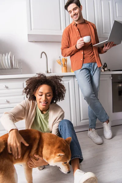 Alegre mujer afroamericana acariciando shiba inu cerca de novio con portátil y café en la cocina - foto de stock