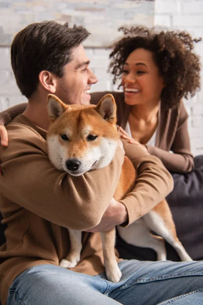 Sonriente hombre abrazando shiba inu perro cerca africano americano novia en casa - foto de stock