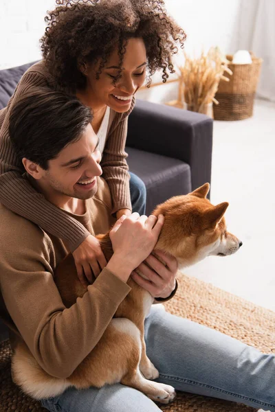 Lächelnde Afroamerikanerin streichelt Shiba Inu in der Nähe ihres Freundes im Wohnzimmer — Stockfoto