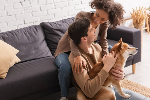 Heureux couple interracial caressant shiba inu et se regardant à la maison — Photo de stock