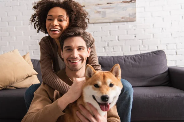 Positive multiethnic couple looking at camera near blurred shiba inu in living room — Stock Photo