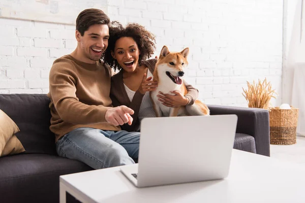 Hombre positivo apuntando a la computadora portátil borrosa cerca de novia afroamericana con shiba inu en casa - foto de stock