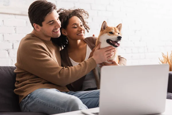 Casal multiétnico tocando shiba inu perto de laptop borrado em casa — Fotografia de Stock