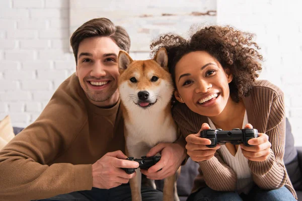 KYIV, UCRANIA - 22 de diciembre de 2021: Pareja multiétnica jugando videojuegos cerca del perro shiba inu en casa - foto de stock