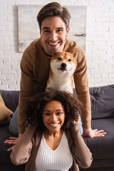 Positiva pareja interracial mirando a la cámara cerca de shiba inu perro en el sofá en casa - foto de stock