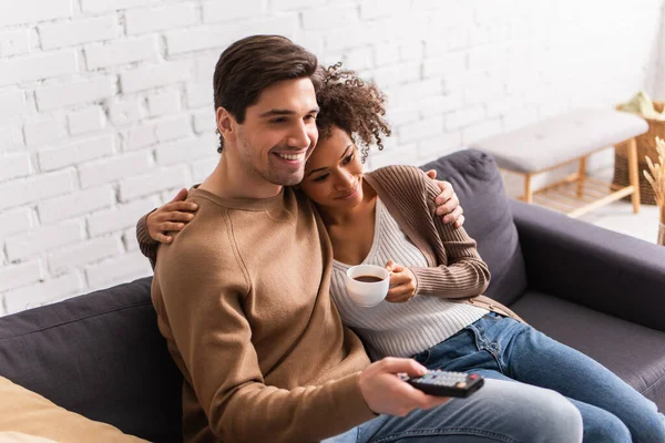 Sorridente donna afroamericana con caffè abbracciando fidanzato con telecomando a casa — Foto stock