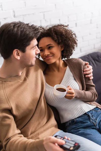 Casal interracial com café e controle remoto olhando um para o outro em casa — Fotografia de Stock