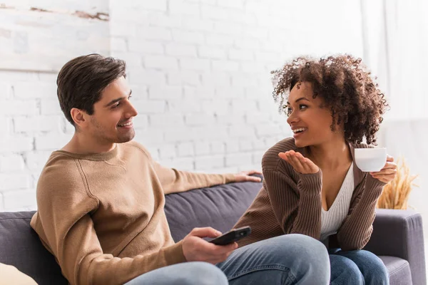 Smiling african american woman with cup talking to boyfriend with remote controller at home — Stock Photo