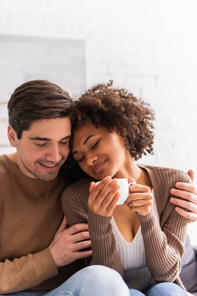 Homem abraçando afro-americana namorada com xícara de café em casa — Fotografia de Stock