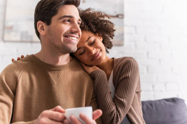 Sonriente mujer afroamericana abrazando novio con taza borrosa en casa - foto de stock