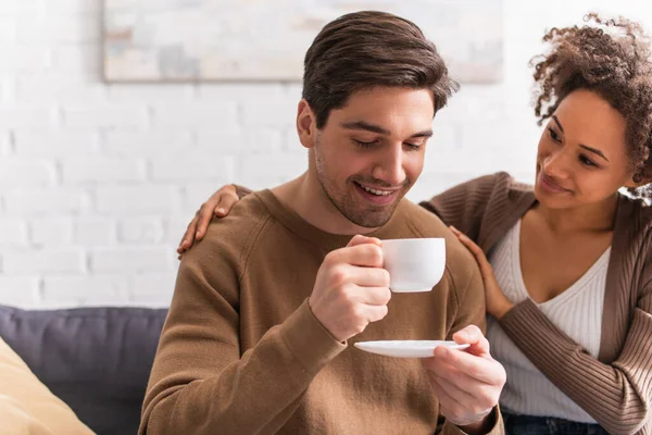 Sorridente donna afro-americana che abbraccia il fidanzato con una tazza di caffè in soggiorno — Foto stock