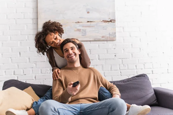 Lächelnder Mann vor dem Fernseher in der Nähe der afrikanisch-amerikanischen Freundin auf der Couch im Wohnzimmer — Stockfoto