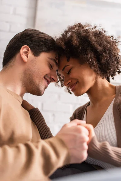 Pareja multiétnica con los ojos cerrados cogidos de la mano en casa - foto de stock