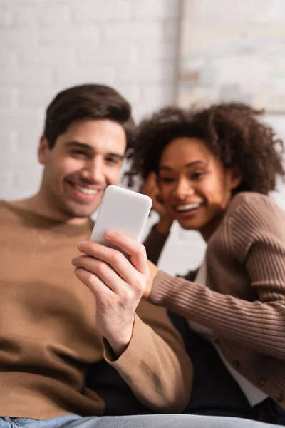 Smartphone in hand of blurred man near african american girlfriend at home — Stock Photo