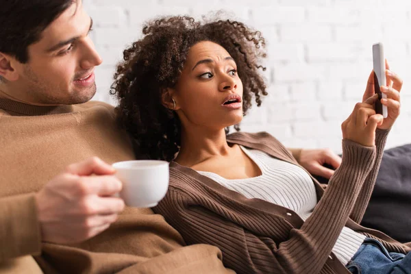 Mujer afroamericana apuntando a teléfono inteligente cerca de novio con taza borrosa en casa - foto de stock