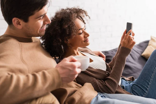Sonriente pareja interracial con café usando teléfono móvil en casa - foto de stock
