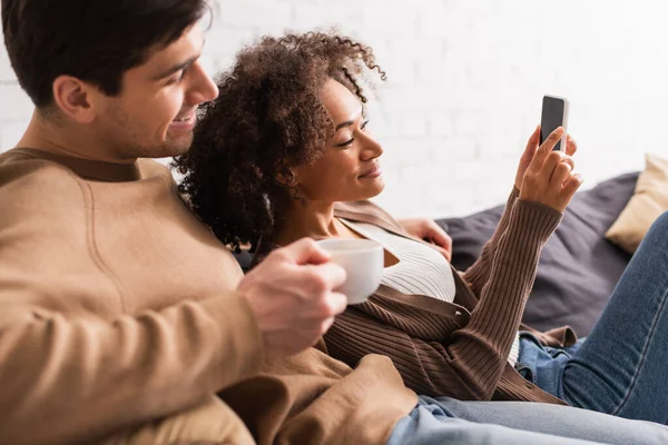 Lächeln gemischtrassiges Paar mit Kaffee mit Handy zu Hause — Stockfoto