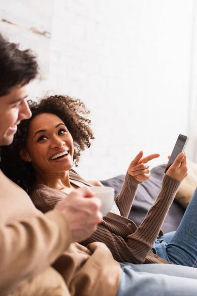 Sourire femme afro-américaine pointant vers smartphone près de petit ami avec tasse floue dans le salon — Photo de stock