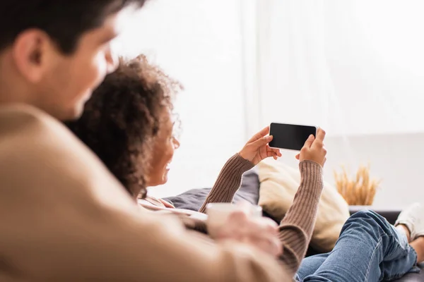 Mujer afroamericana sosteniendo teléfono inteligente cerca de novio borroso con taza en casa - foto de stock