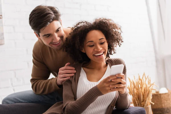 Sorrindo homem abraçando afro-americana namorada usando smartphone em casa — Fotografia de Stock