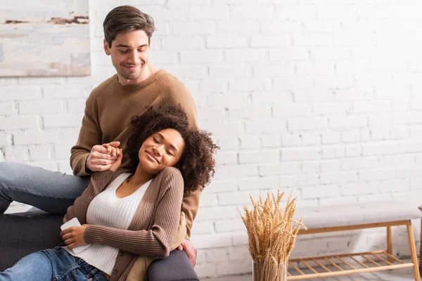 Positive interracial couple with cellphone holding hands at home — Stock Photo