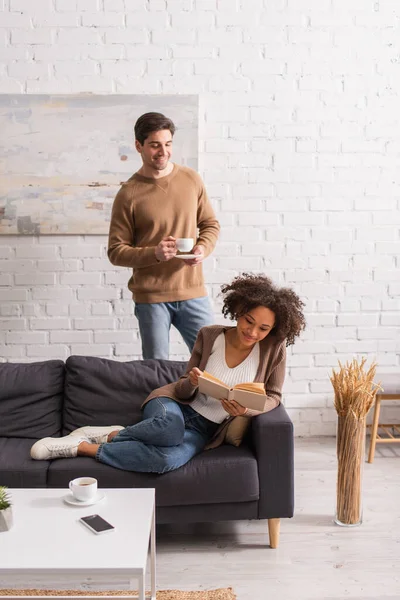 Man holding coffee cup near african american girlfriend reading book on couch at home — Stock Photo