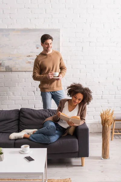 Hombre sosteniendo taza de café cerca de novia afroamericana leyendo libro en el sofá en casa - foto de stock