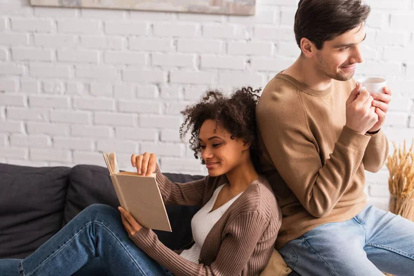 Man holding cup of coffee while african american girlfriend reading book at home — Stock Photo