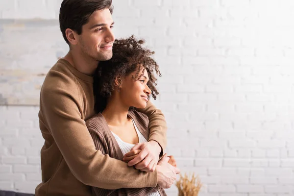 Sonriente hombre abrazando rizado africano americano novia en casa - foto de stock