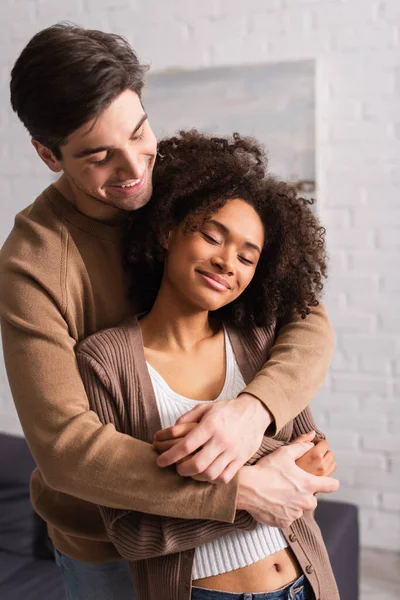 Homem positivo abraçando a namorada afro-americana em casa — Fotografia de Stock