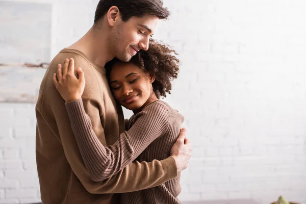 Sonriente pareja multiétnica abrazándose en la sala de estar - foto de stock