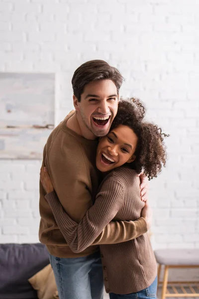 Positiva pareja multiétnica abrazando y mirando a la cámara en casa - foto de stock