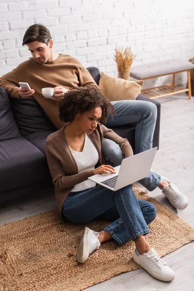 Afro-Américain pigiste en utilisant un ordinateur portable près du petit ami avec café et smartphone sur le canapé à la maison — Photo de stock