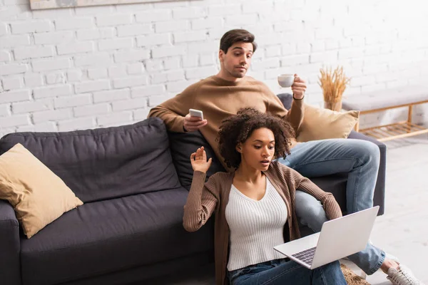 Libero professionista afroamericano che parla con fidanzato vicino al fidanzato con caffè e cellulare a casa — Foto stock