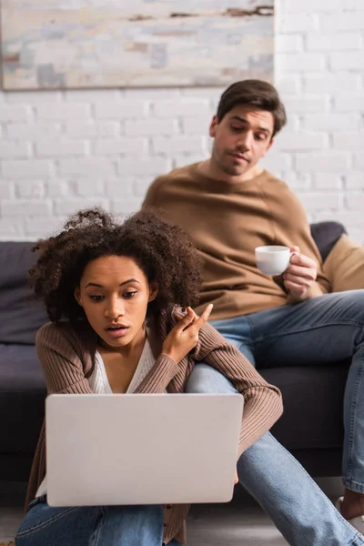 Freelancer afroamericano con laptop apuntando con el dedo al novio borroso con café en casa - foto de stock