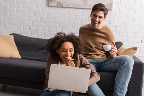 Felice freelance africano americano utilizzando laptop vicino fidanzato con tazza sul divano — Foto stock