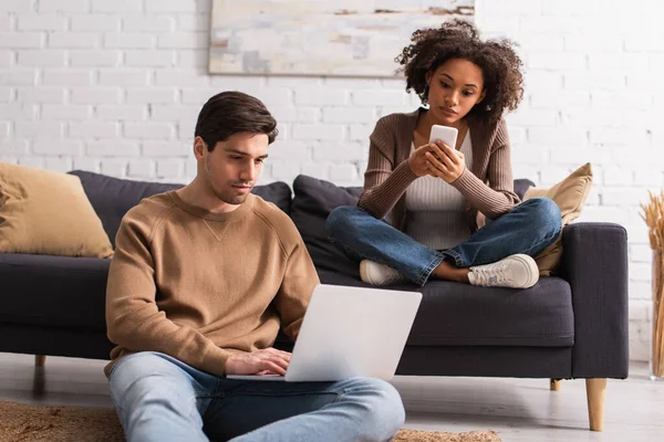 Freelancer usando laptop cerca de novia afroamericana con celular en casa - foto de stock