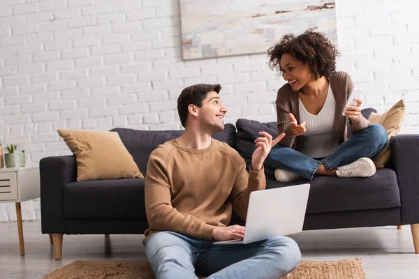 Sorrindo casal interracial com dispositivos apontando com os dedos em casa — Fotografia de Stock